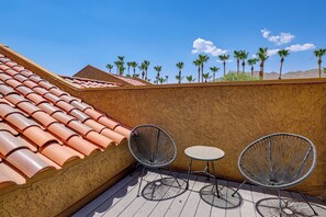 Eine Dachterrasse mit Terrakottafliesen, zwei Metallstühlen, einem kleinen runden Tisch und Blick auf Palmen vor blauem Himmel.