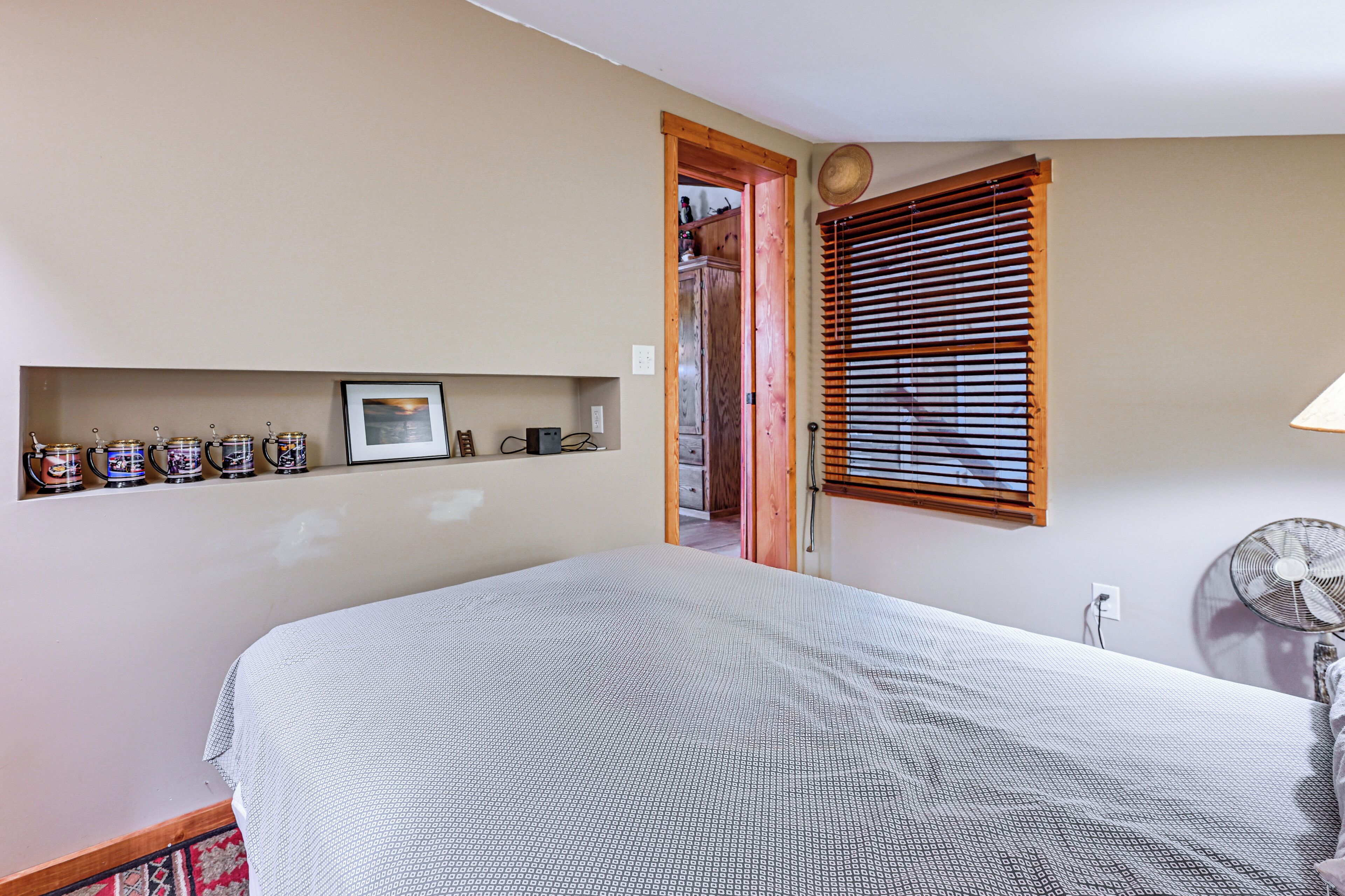 Une chambre à coucher avec un lit, une étagère contenant des ustensiles de cuisine, un ventilateur et une fenêtre avec des stores.