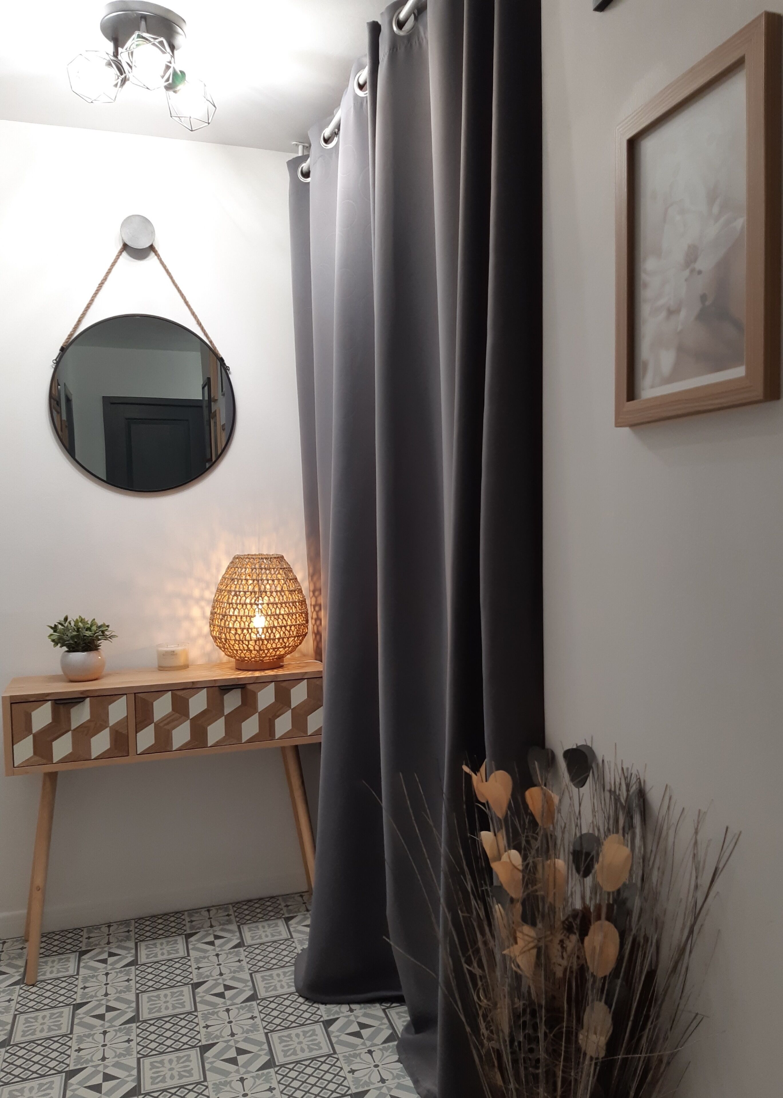 Une pièce avec un miroir rond, une console en bois ornée d’un motif géométrique, un lampadaire, un rideau foncé et un tableau encadré accroché au mur.