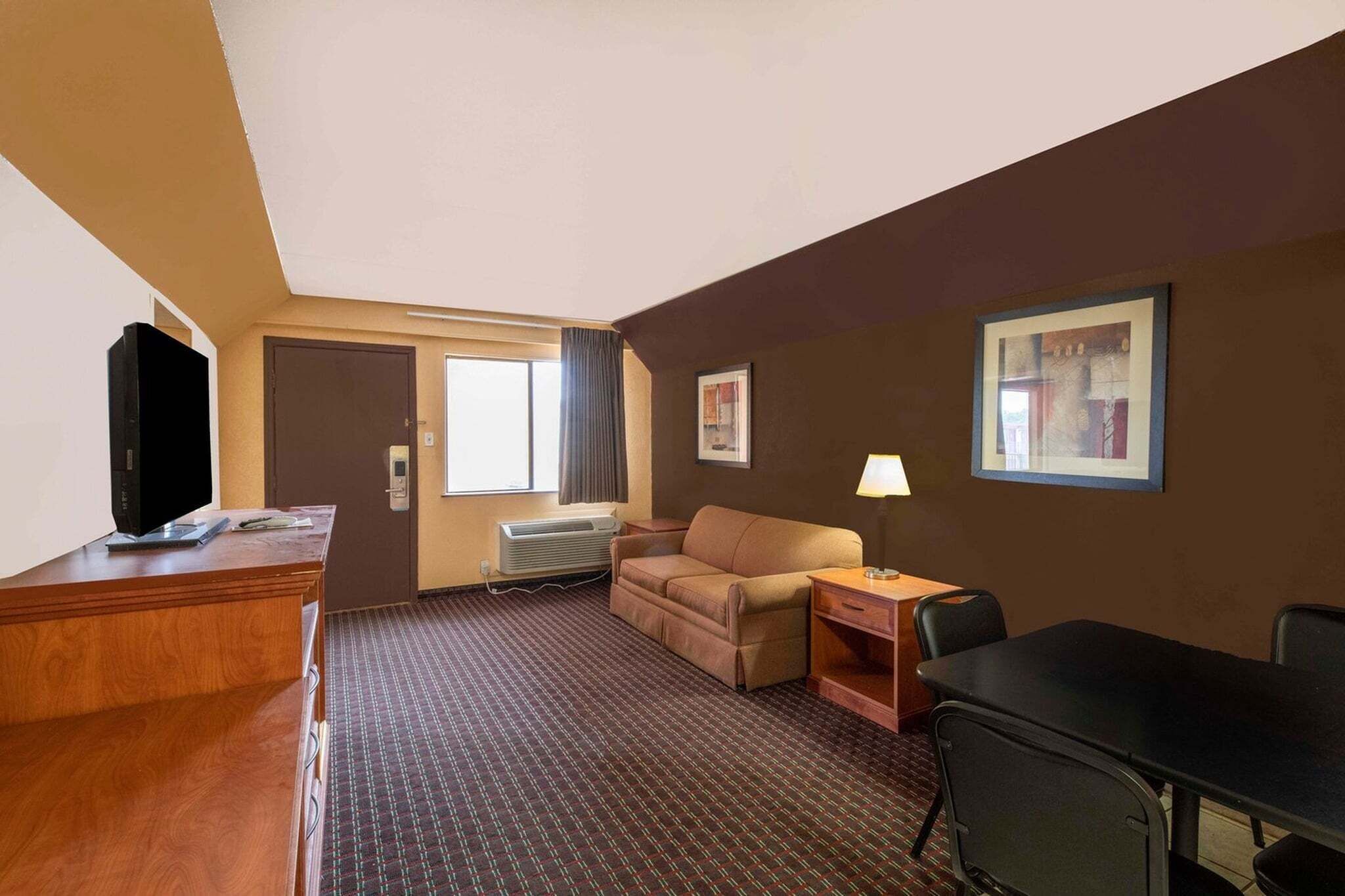 A hotel room with a wooden desk, a television, a sofa, a small table with a lamp, and a window with curtains.