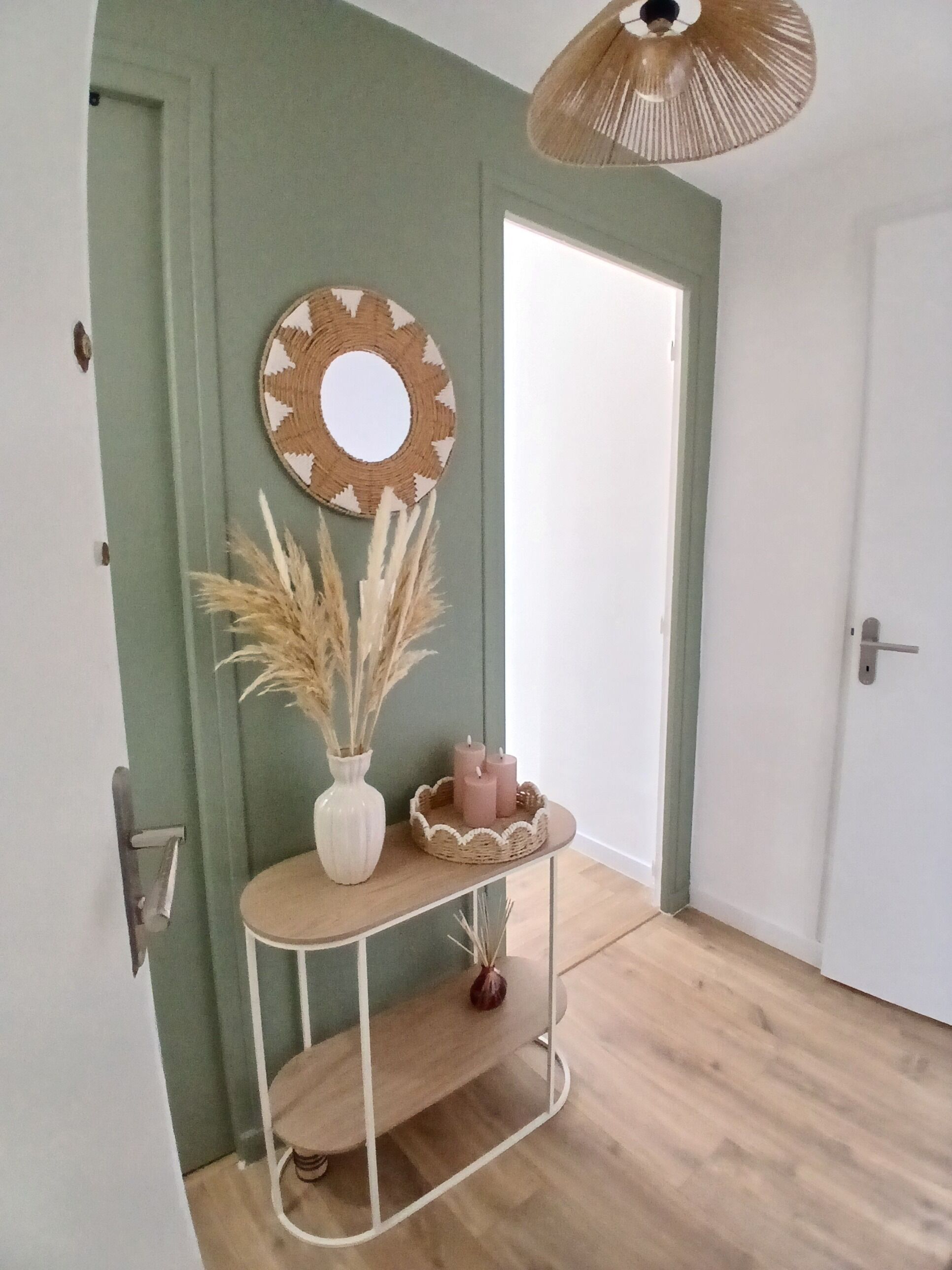 Une pièce avec un mur vert, un miroir rond, une petite table blanche avec des bougies et un panier en osier.