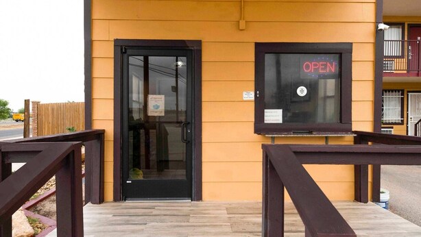 Un bâtiment avec une porte noire, une fenêtre sur laquelle est affiché un panneau indiquant « OUVERT », et une terrasse en bois dotée de rampes.