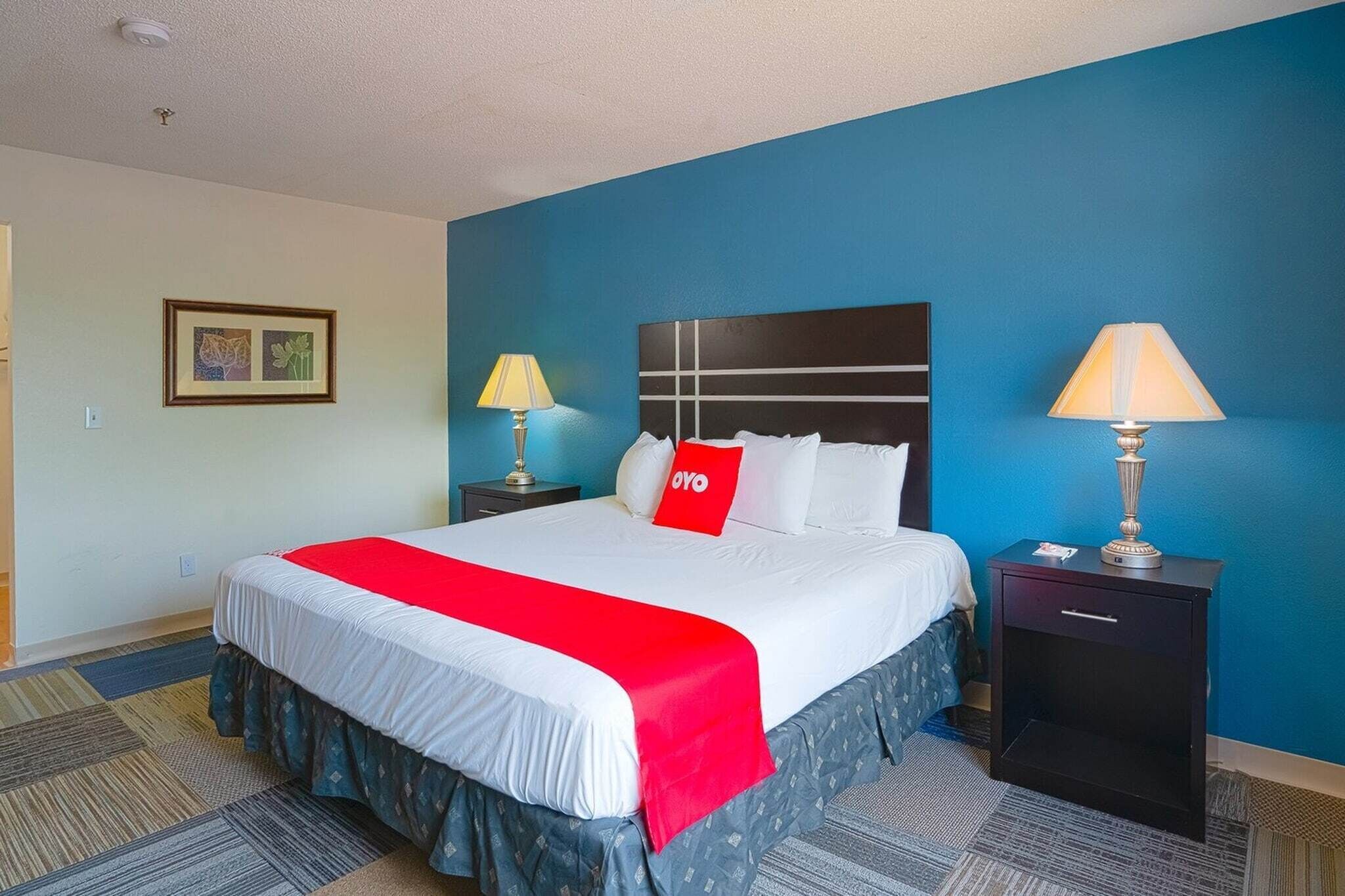 A hotel room with a bed, two bedside tables with lamps, a red throw blanket, and a framed picture on the wall.