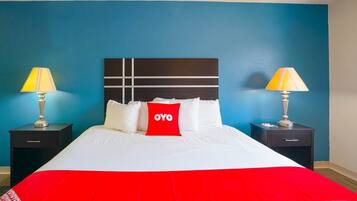 Ein Hotelzimmer mit einem großen Bett, zwei Nachttischen mit Lampen, einem roten Kopfkissen mit 'OYO'-Branding und einer blauen Wand.