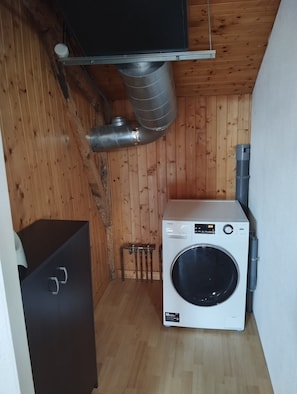 Une petite buanderie avec un lave-linge blanc, une grille de ventilation en métal et un plancher en bois.