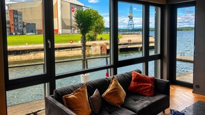 A modern room with large windows, a black leather sofa, and a view of a water body and industrial buildings.