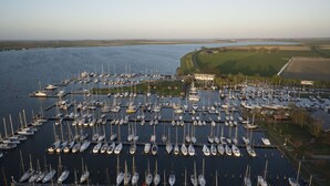 A marina with numerous boats docked, a building with a green roof, and a view of the water and fields in the background. - Stay on a luxury houseboat at Lake Veerse in Wolphaartsdijk. Enjoy a rooftop terrace, private mooring, water sports and Zeeland's nature. (Wolphaartsdijk)
