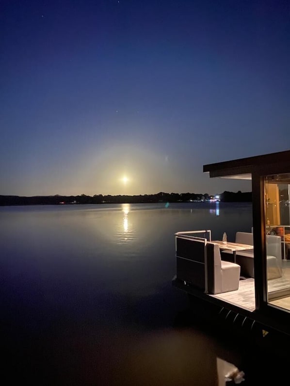Une vue paisible sur le lac au crépuscule, avec un espace repas comprenant des sièges extérieurs et une véranda en verre.