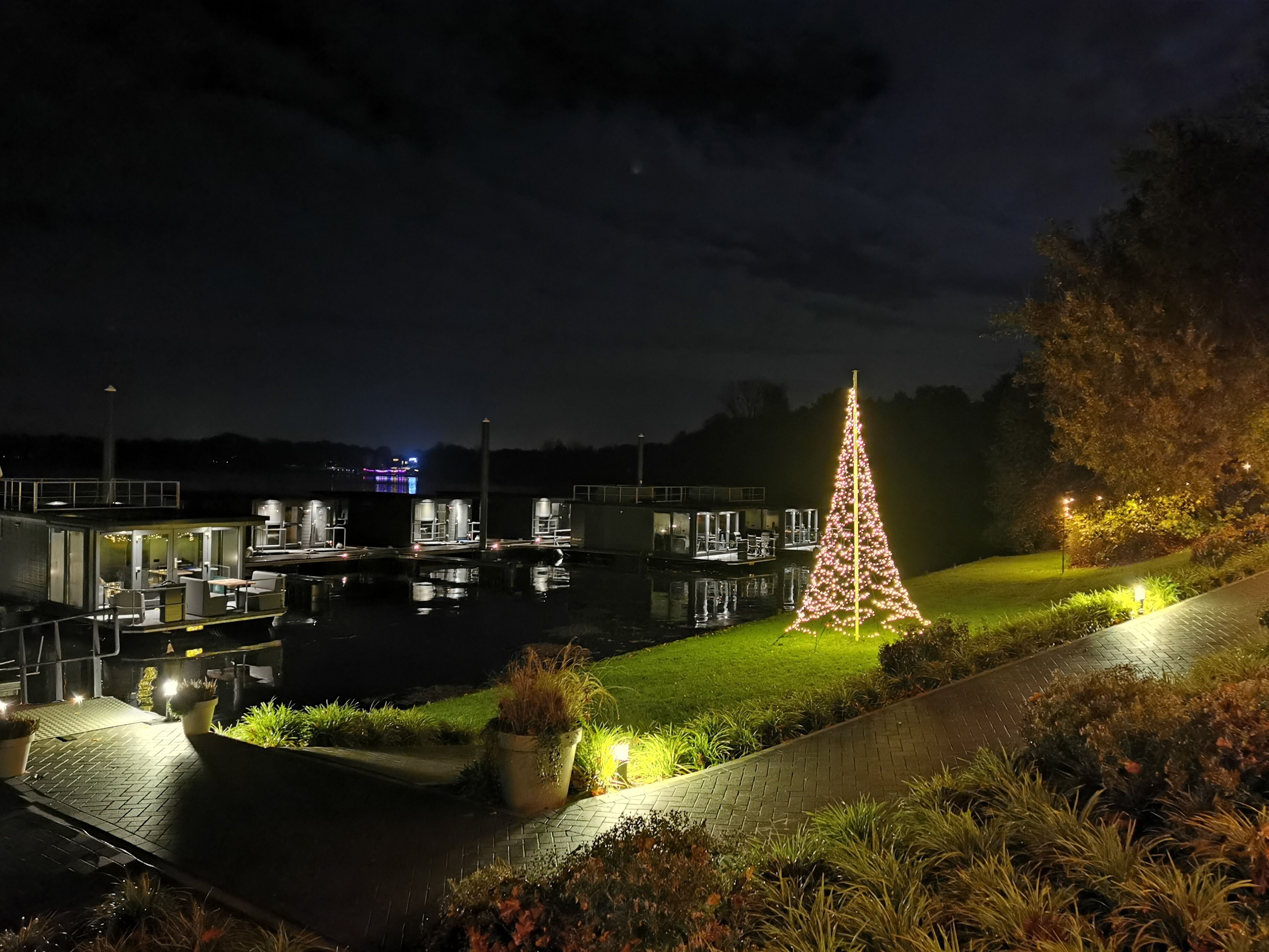 A waterfront property with a large illuminated Christmas tree, a dock with boats, and a building with multiple levels and outdoor seating.