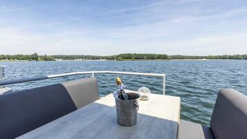 A boat with a table and chairs, a metal bucket with a bottle, and a view of a lake with a distant shoreline.