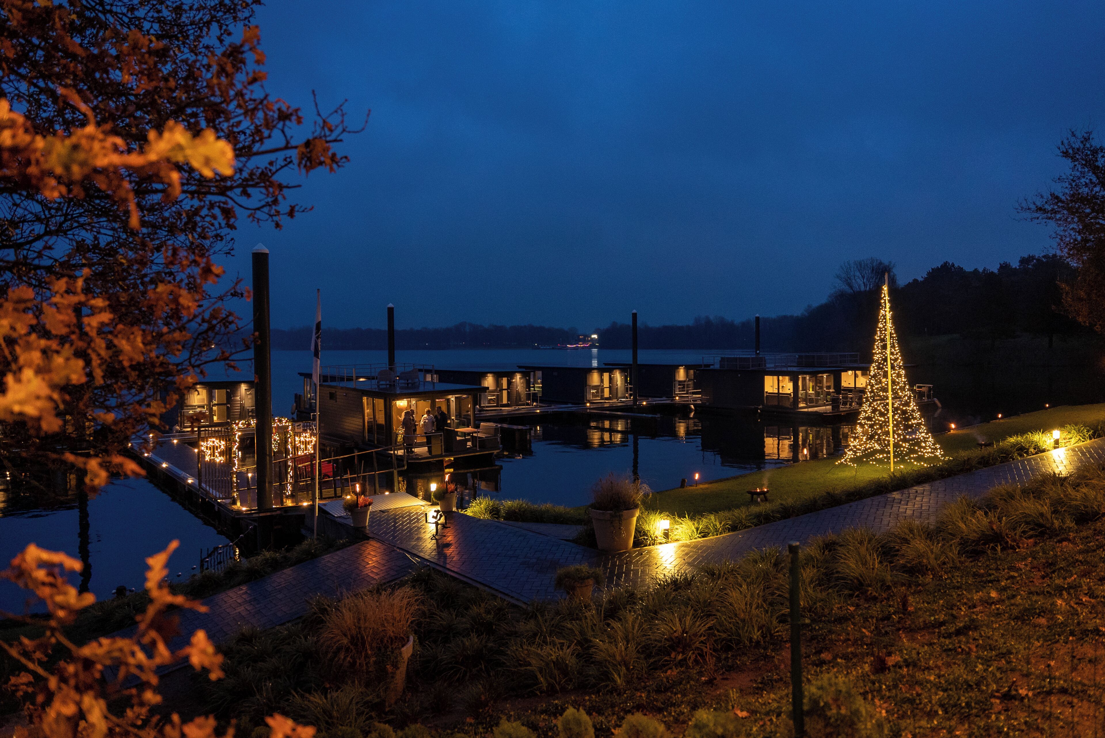 Een pand aan het water met een verlichte kerstboom, een steiger en terrasjes.