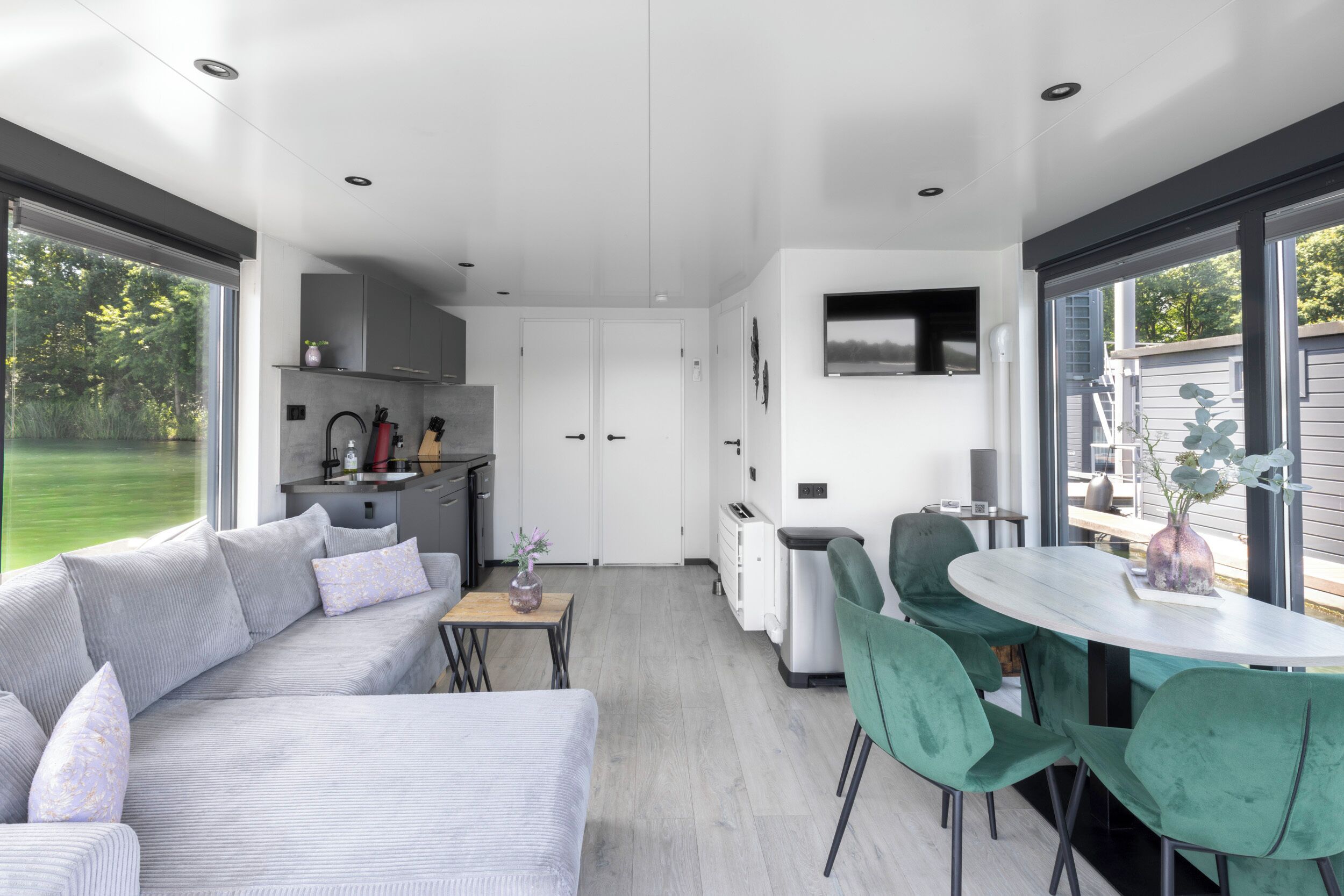 A modern kitchen with a built-in oven, a dining area with a round table and green chairs, and a large window overlooking a garden.
