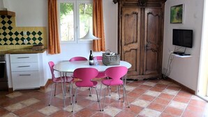 Un comedor con una mesa blanca y sillas rosadas, un armario de madera, un televisor sobre un mueble bajo y una ventana con cortinas.
