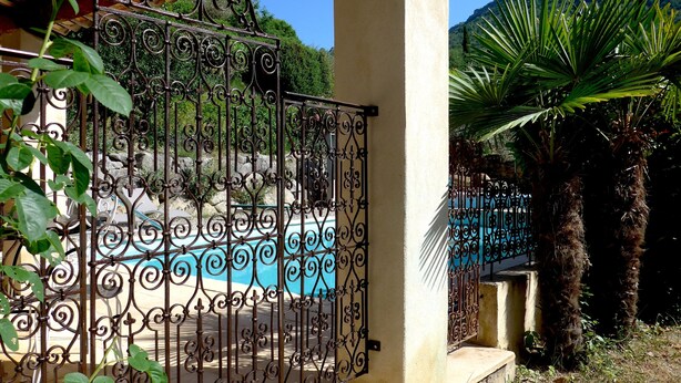 Una entrada con reja, piscina, palmeras y montañas al fondo.