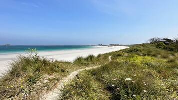 Un sentier qui longe la plage et mène à une étendue de sable fin, bordée d’une eau d’un bleu limpide et offrant une vue sur des reliefs lointains.
