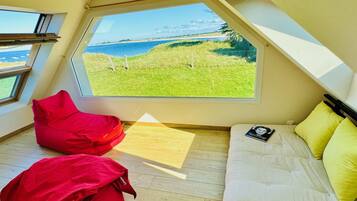 Une pièce avec une grande fenêtre, un lit, un pouf rouge et un plancher en bois.