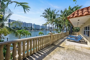 A balcony with a view of a body of water, palm trees, and a building with a tiled roof.