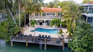 A two-story building with a swimming pool, palm trees, and a dock extending into a body of water.