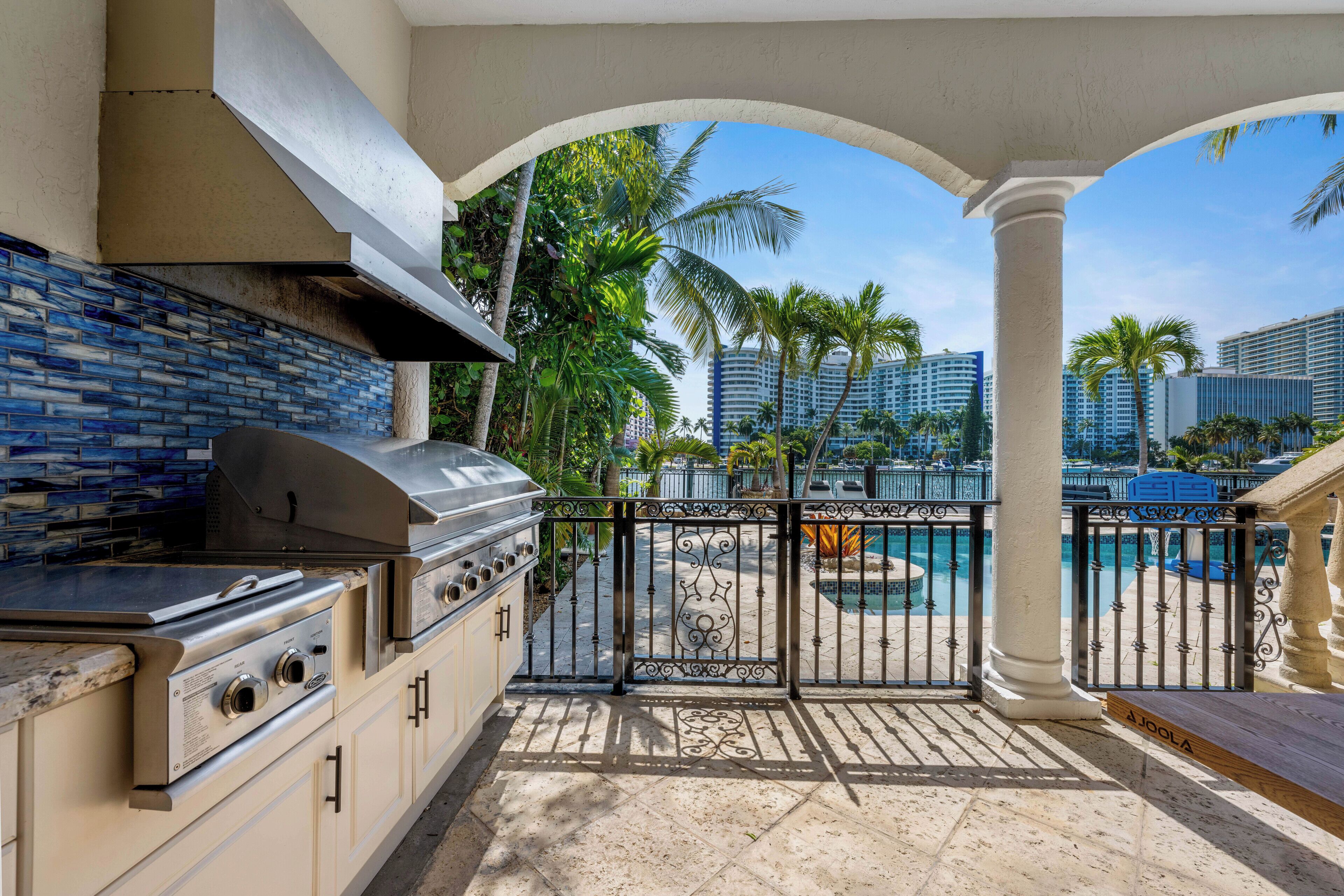 An outdoor kitchen with a grill, a view of a swimming pool, and palm trees.