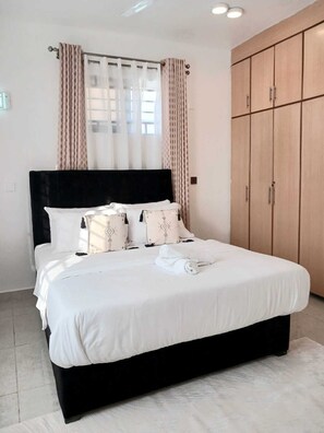 A neatly made bed with white bedding and decorative pillows, a black headboard, and a large wardrobe with multiple doors. - Shamm dream riverside (Nairobi)