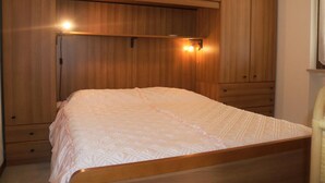 Quarto com estrutura de cama em madeira, colcha acolchoada branca e armários embutidos em madeira, com uma prateleira sobre a cama.