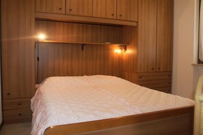 Een slaapkamer met een houten bedframe, een wit geborduurde spreidek en ingebouwde houten kasten met een plank boven het bed.