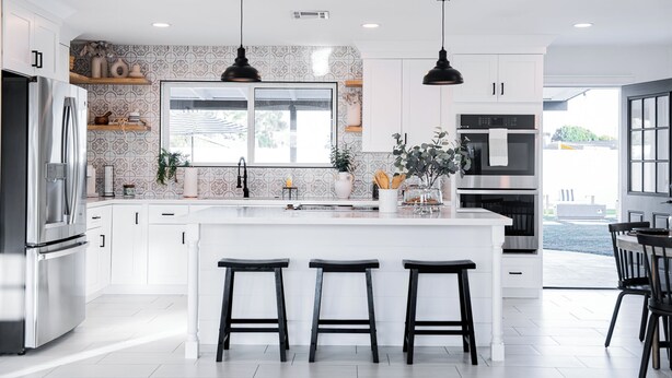 Una cocina moderna con armarios blancos, un refrigerador de acero inoxidable, una isla grande con taburetes de bar, iluminación colgante y vistas al exterior.