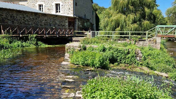 Un bâtiment en pierre avec un pont métallique enjambant un ruisseau, le tout entouré de verdure.