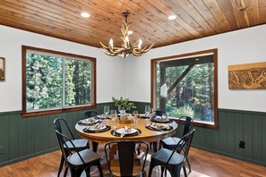 Ein Esszimmer mit rundem Tisch, Stühlen, einer Chandelieber und großen Fenstern mit Blick auf einen Wald.