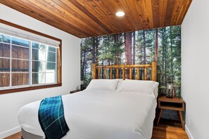 Una camera da letto con soffitto in legno, un letto con biancheria da letto bianca, una coperta a quadri blu e bianchi, un comodino con una lanterna e carta da parati con motivo foresta.