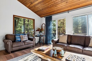 A living room with a wooden ceiling, two brown leather sofas, a coffee table, and large windows with a view of trees. - Pine-View Big Bear Cabin • Hot Tub + Game Room | London Pines Lodge (Big Bear)