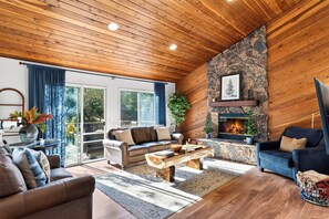 Ein gemütliches Wohnzimmer mit Kamin aus Stein, Holzboden und einem großen Fenster mit Vorhängen.