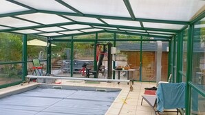 A conservatory with a swimming pool, large windows, and various items scattered around.