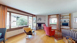 Een ruime woonkamer met grote ramen, een open haard, een bank, een fauteuil en een boekenkast.