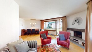 Ein Wohnzimmer mit Kamin, roten Armlehnstühlen, einem gestreiften Sofa, einem Essbereich mit Klavier und einem großen Fenster mit Vorhängen.