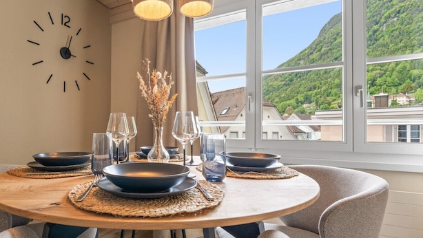 Un coin repas avec une table ronde, des assiettes noires, des verres transparents et une horloge au mur.