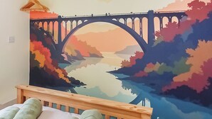 Una cama con cabecera de madera, un mural colorido de un puente y paisaje, y toallas verdes sobre la cama.