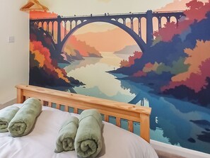 Ein Bett mit einem Holz-Kopfstück, einer farbenfrohen Wandmalerei einer Brücke und Landschaft, und grünen Handtüchern auf dem Bett.