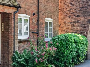 A brick building with two white-framed windows, a black pipe on the wall, and a green hedge with pink flowers.