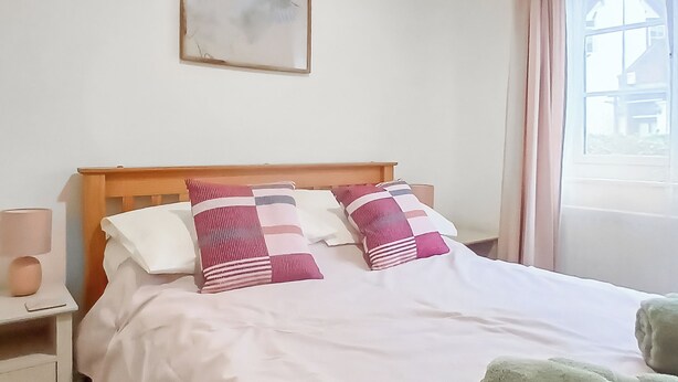 Una cama bien tendida con ropa de cama blanca y dos almohadas decorativas, una lámpara de noche y una ventana con cortinas.