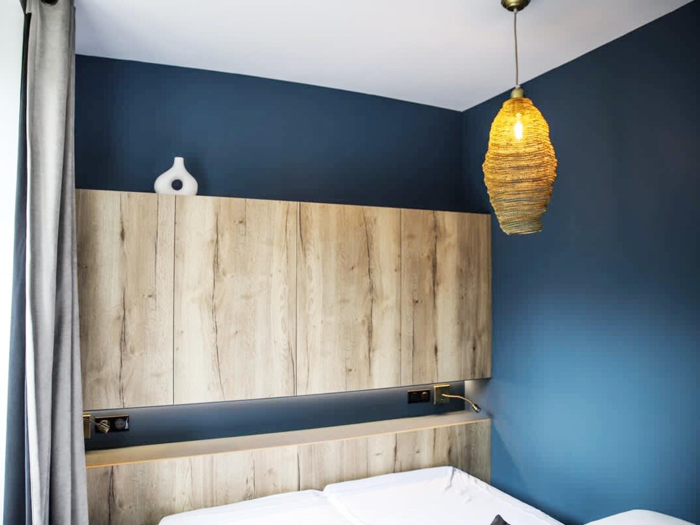 A bedroom with a wooden headboard, a bedside table, a hanging light fixture, and a wall-mounted shelf.