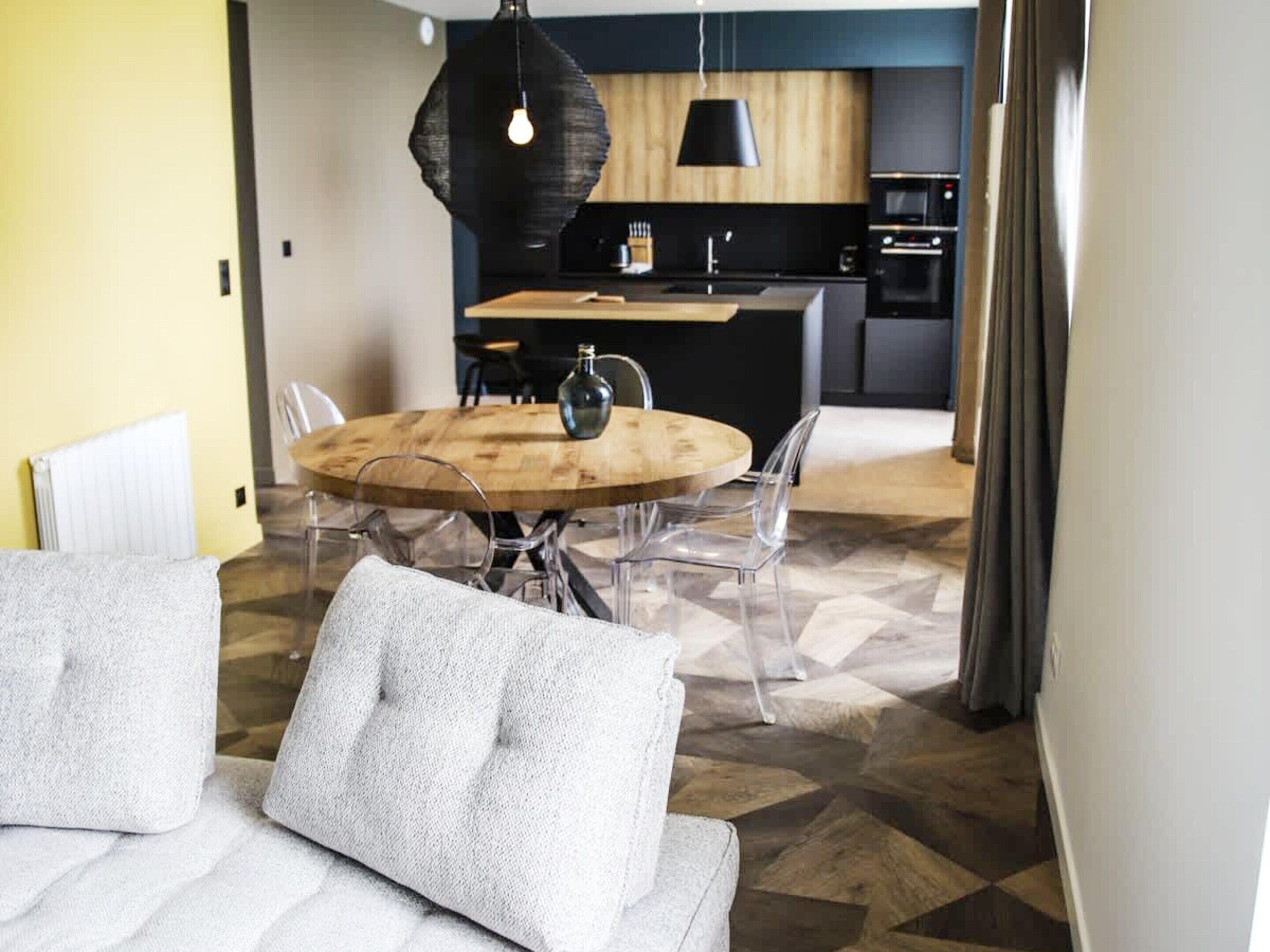 A modern kitchen with a round wooden table, clear chairs, and a hanging light fixture.