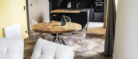 A modern kitchen with a round wooden table, clear chairs, and a hanging light fixture.