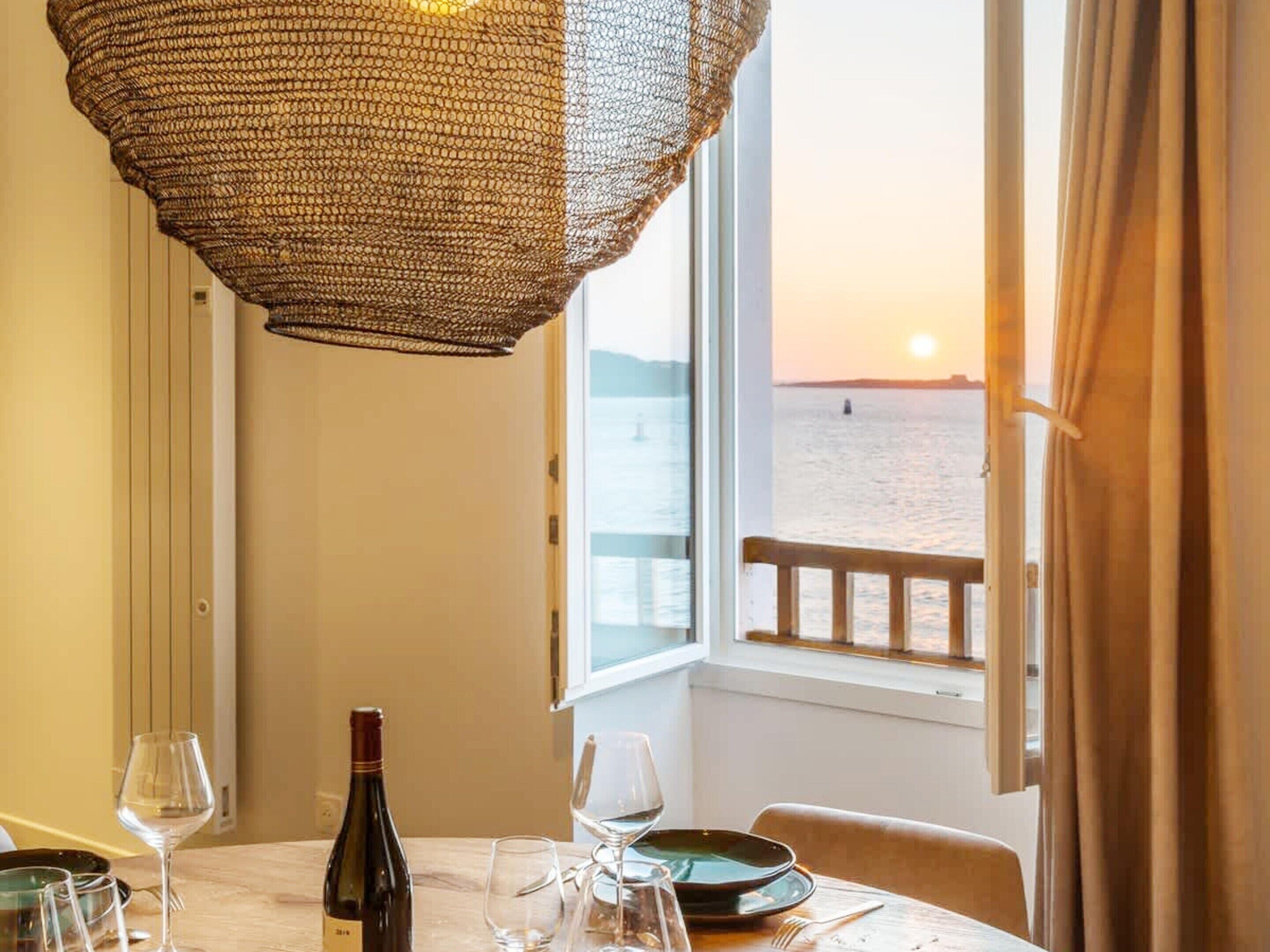 A table set for dining with a bottle of wine, glasses, and plates, overlooking a body of water at sunset.
