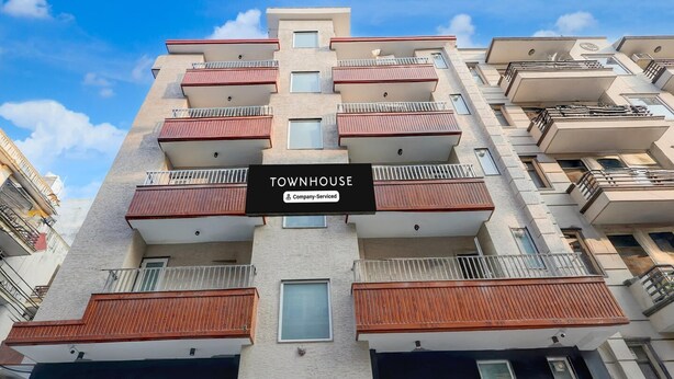 En byggnad med flera våningar och balkonger, en skylt med texten ”TOWNHOUSE – företagsservice” och en klar himmel.