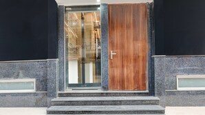 En modern entré till en byggnad med en glasdörr och en trädörr, omgiven av mörka stenmurar och trappsteg som leder upp till entrén.