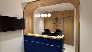 A reception desk with a blue counter, a flat-screen TV mounted on the wall, and a wooden-framed archway leading to a room with a sign reading 'The Shivanta Inn'. - The Shivanta Inn (Gandhinagar)