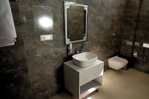 A bathroom with a marble wall, a white sink, a toilet, and a shower. - Hotel Maurya Greens (Purnagiri)
