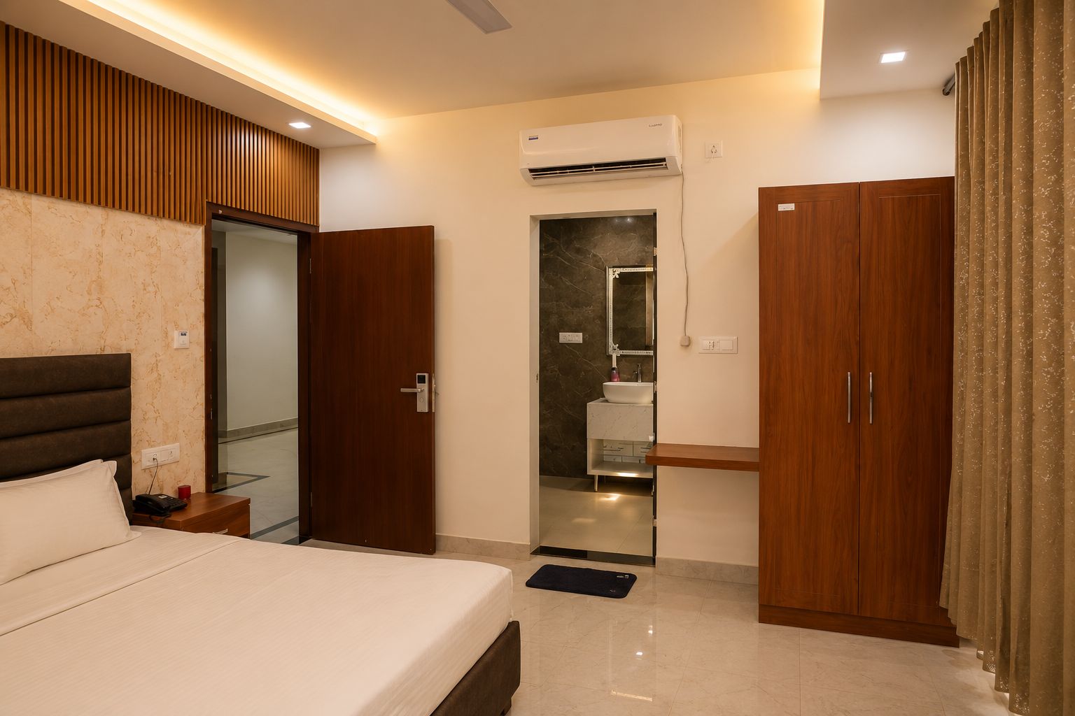 A hotel room with a bed, a bedside table, a wardrobe, an air conditioning unit, and a bathroom with a mirror and sink.