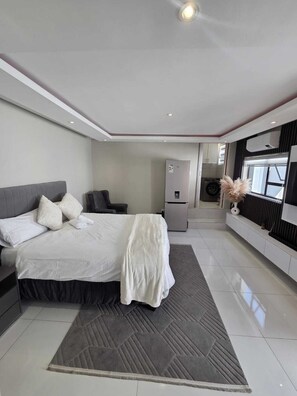 A modern bedroom with a bed, a desk, a chair, a refrigerator, and a window. - Private & Modern Studio Apartment in Sandton (Johannesburg)